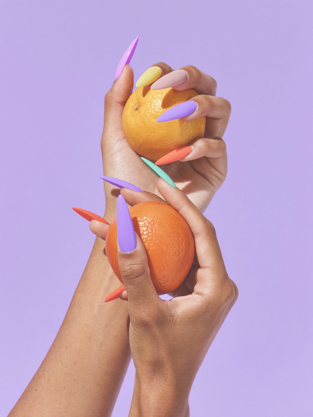 Hands with pastel rainbow nails holding an orange on a lilac backdrop